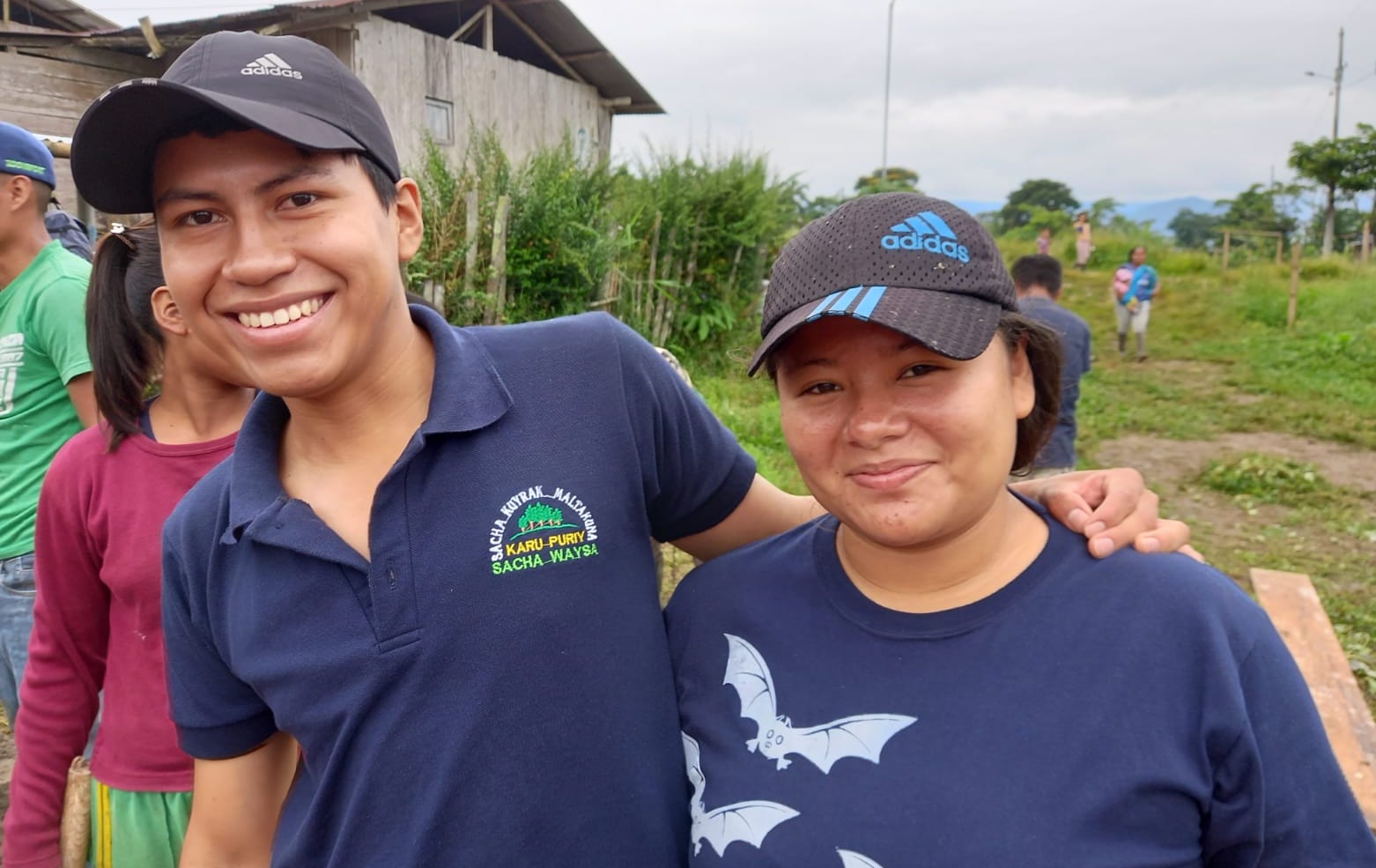 María and Jimmy from Sacha Kuyrana Maltakuna are stood together, smiling. They are wearing baseball caps and blue tops, and stood in a green area.